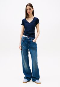 Žena v tmavě modrém tričku s výstřihem do V, volných modrých džínách, světle modrých teniskách a s viditelným páskem s logem, stojící na bílém pozadí.