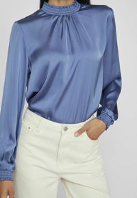Satinbluse in sanftem Blau mit gefaltetem Stehkragen und langen Ärmeln; geraffte Manschetten. Kombiniert mit cremefarbener Denim-Jeans mit Knopfverschluss.