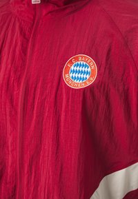 Giacca rossa con zip realizzata in materiale leggero, dotata di una patch del logo del Bayern Monaco e una striscia bianca lungo l'orlo.