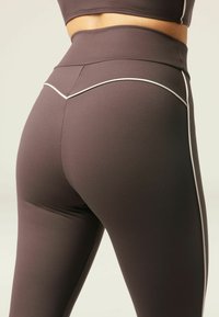 Brown high-waisted leggings with white piping detail along the side seams and a smooth, stretchy texture, designed for comfort and flexibility.