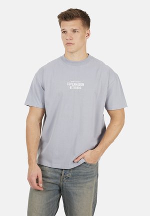 Młody mężczyzna ubrany w jasnoszarą koszulkę z napisem "Copenhagen Østerbro" i współrzędnymi, w połączeniu z wyblakłymi niebieskimi dżinsami, stoi z jedną ręką w kieszeni.