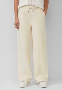 Person, die eine cremefarbene weite Sweatpants mit Kordelzug und weiße Plateausneaker trägt, steht vor einem grauen Hintergrund.