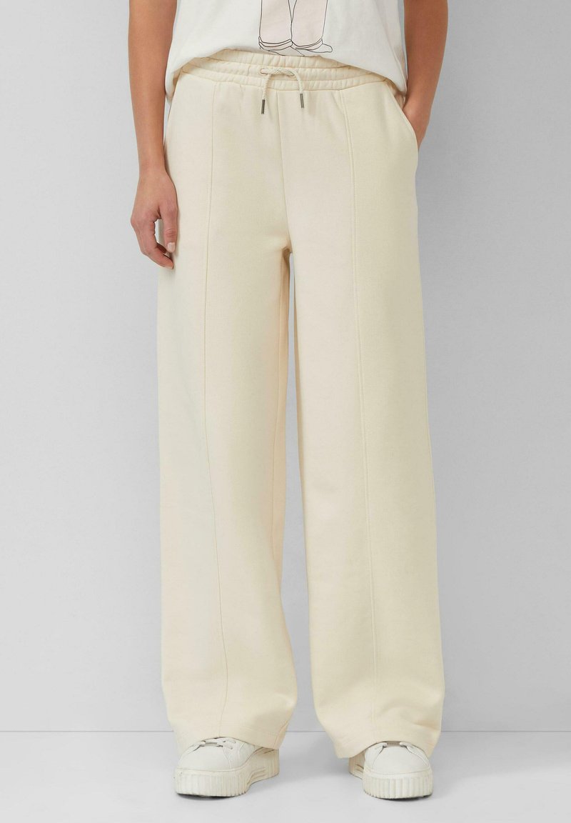 Person, die eine cremefarbene weite Sweatpants mit Kordelzug und weiße Plateausneaker trägt, steht vor einem grauen Hintergrund.