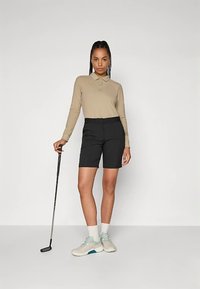 Beżowa koszulka polo z długim rękawem, czarne dopasowane szorty, kij golfowy w ręku oraz jasne buty z turkusowymi akcentami. Prosty, nowoczesny strój sportowy.