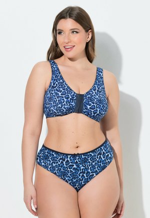 Donna che indossa un reggiseno stampa leopardo blu e nero e mutandine a vita alta abbinate, sorridente e che guarda di lato su sfondo bianco.