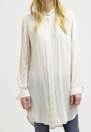 Femme portant une longue chemise blanche boutonnée avec un ourlet arrondi et un jean bleu, debout devant un fond uni.