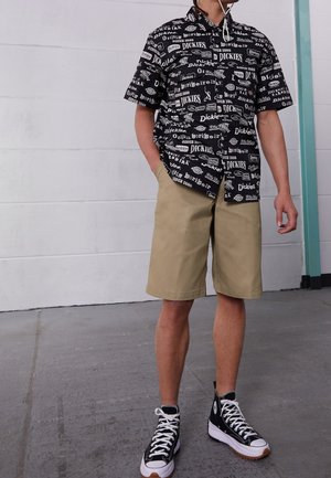 Chemise noire à manches courtes avec logos graphiques blancs, assortie à un short beige. Chaussures montantes noires avec accents blancs.