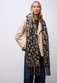 Strikket tørklæde med et sort og beige leopardmønster, med frynser i enderne. Båret med en lys beige jakke og mørke jeans.