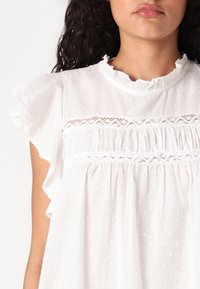 Blouse blanche à manches courtes avec volants, texture à pois, dentelle au niveau de la poitrine et tissu froncé à la taille.