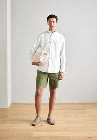 Vit skjorta med knappar, gröna shorts och beige slip-on skor. Håller i ett rullat beige tyg. Släta texturer och avspänd design.