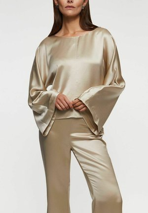 Nainen, joka pukeutunut beigeen satiiniseen pitkähihaiseen puseroon, jossa on leveät kalvosimet, ja vastaaviin satiinisiin housuihin, seisoo kämmenet edessään ristissä.