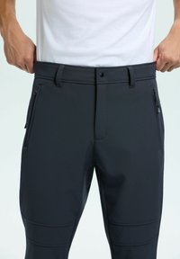 Händer som drar i midjebandet på svarta byxor med dragkedjefickor, som bärs med en vit skjorta, visad från midjan till mitten av låren.