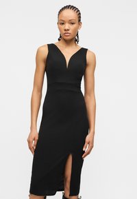 Γυναίκα με πλεξούδες, ντυμένη με μαύρο μίντι φόρεμα χωρίς μανίκια, με V λαιμόκοψη και σ κίσιμο μπροστά, στέκεται μπροστά σε απλό φωτεινό φόντο.