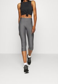 Czarny krótki top z węzłami w połączeniu z szarymi legginsami capri. Oba wykonane z rozciągliwego materiału. Modelka nosi czarne buty sportowe z białymi skarpetkami.