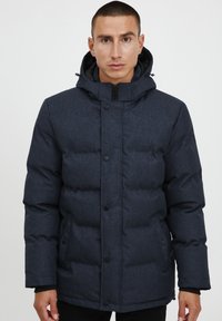 Mörkblå puffjacka med huva, dragkedjestängning och knappdetaljer, med en mjuk texturerad yta och vadderad design för värme.