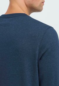 Férfi sötétkék hosszúujjú ingben, hátulról és részben oldalprofilból látható, egy sima világos háttér előtt.