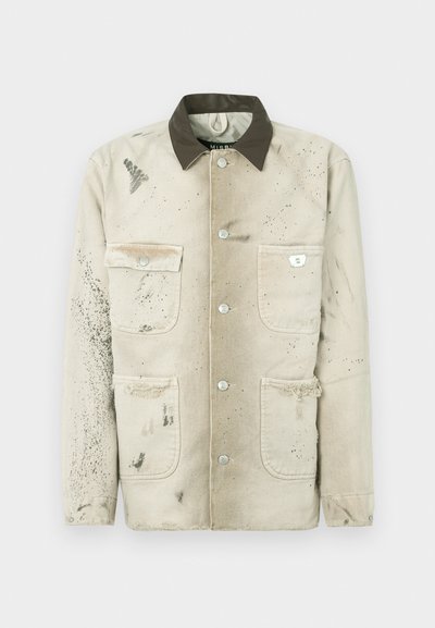 Veste en toile beige avec un col foncé, quatre poches, des boutons en argent et une texture usée avec des éclaboussures de peinture.