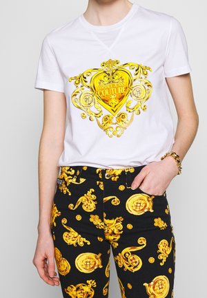 Biała koszula z krótkim rękawem z dużym złotym barokowym sercem i napisem „Versace Jeans Couture”, zestawiona z czarnymi spodniami z złotymi barokowymi wzorami.
