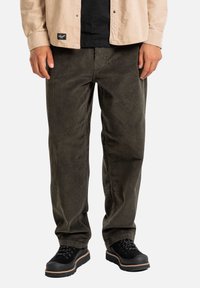 Pantaloni in velluto a coste di colore verde scuro, con gamba dritta e texture a coste, abbinati a scarpe nere stringate e una camicia beige con bottoni.