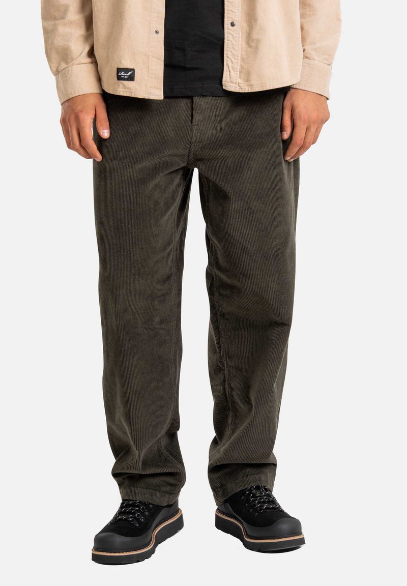 Pantaloni in velluto a coste di colore verde scuro, con gamba dritta e texture a coste, abbinati a scarpe nere stringate e una camicia beige con bottoni.