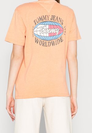 Osoba nosząca brzoskwiniową koszulkę z logo globu oraz napisem "Tommy Jeans Worldwide Since 1985" na plecach, w połączeniu z białymi spodniami.