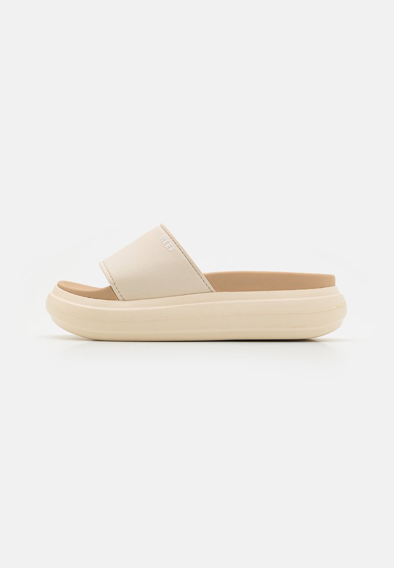 Sandal de slide bege com uma faixa larga em tecido e uma sola almofadada de cor creme. Textura suave com design minimalista.