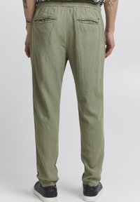Pantalon en lin vert olive avec une coupe décontractée, doté de deux poches arrière et d'une taille élastique. Porté avec des baskets noires.