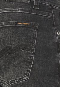 Black denim jeans back pocket with black stitching detail and small orange "Nudie Jeans Co" label above the pocket.