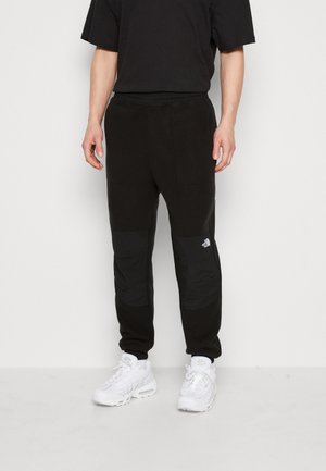 Mann trägt schwarze Jogginghosen mit verstärkten Kniepolstern und weißen Sneakers, steht vor einem schlichten hellen Hintergrund.
