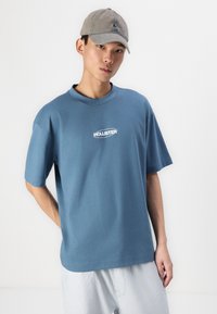 Kék pamut póló, kerek nyakú és rövidujjú, a mellkason fehér "Hollister" logóval. Az egyszerű kialakítás minta nélküli.