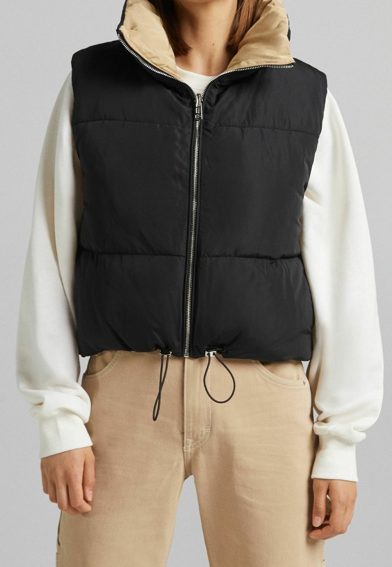 Black padded vest with a beige inner lining, zip closure, and adjustable black drawstrings at the hem. Worn over a white long-sleeve shirt.