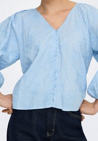 Femme portant une blouse bleu clair boutonnée avec des motifs floraux brodés et un jean en denim bleu foncé, mains posées sur les hanches.