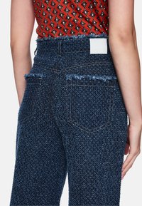 Dunkelblaue, strukturierte Jeans mit fransigem Saum und Gesäßtaschen, kombiniert mit einem gemusterten roten Oberteil. Weiße Markenetikette am Bund sichtbar.