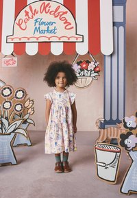 Noor tüdruk lillelise kleidiga seisab värvikas lille-turukarbis, millel on käsitsi joonistatud lilled ja triibuline varikatus.