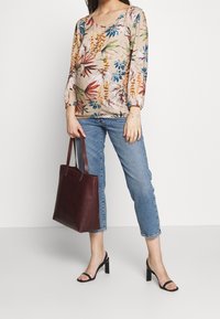 Mujer vestida con una blusa de manga larga floral, jeans ajustados azul cropped, sandalias de tacón negras, sosteniendo un bolso tote de cuero marrón oscuro frente a un fondo liso.