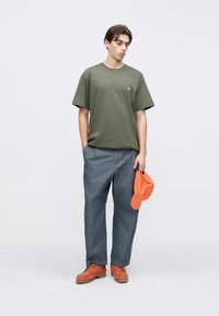 Λαδί βαμβακερό μπλουζάκι, άνετη εφαρμογή· μπλε τζιν παντελόνια· πορτοκαλί καπέλο κρατημένο στο αριστερό χέρι· καφέ παπούτσια με υφή στη σόλα. Απλό, καθημερινό ντύσιμο.