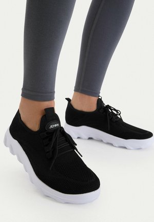 Persona che indossa sneakers in maglia a rete nere con suole bianche e lacci neri, abbinate a leggings grigio scuro, in piedi su una superficie bianca.