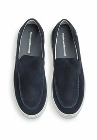 Mockasiner i mocka i marinblått med vita gummisulor. Har kontrasterande sömmar och logotyp på innersulan. Slät textur, rundad form.