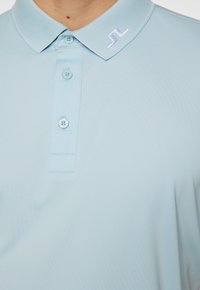 Ljusblå pikétröja med ribbat krage, tre knappar och en liten broderad logotyp i kragen. Slät, lätt strukturerad tyg.