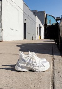 Paire de baskets blanches épaisses avec des lacets sur un trottoir en béton à côté d'une voiture garée avec une porte ouverte sous un ciel bleu clair.