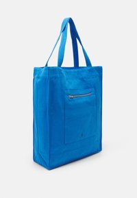 Bolsa de tote azul hecha de lona, con dos asas para el hombro, forma rectangular y un bolsillo frontal con cremallera. Textura suave y color sólido.