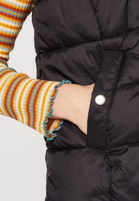 Zwarte puffer vest met een gelamineerde textuur en zilveren drukknoop, gecombineerd met een multicolor gestreepte ribgebreide lange mouw met volants aan de manchetten.