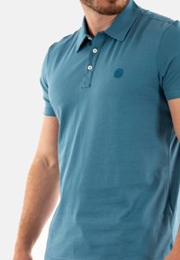 Polo a maniche corte in cotone teal con patta a tre bottoni e logo ricamato sul petto; texture liscia e design aderente.