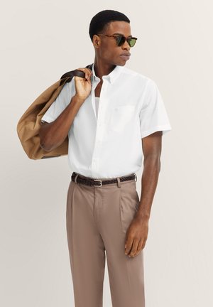 Weißes Kurzarmhemd mit einer Brusttasche, kombiniert mit hellbraunen Hosen und einem geflochtenen Gürtel. Braune Jacke über der Schulter drapiert.