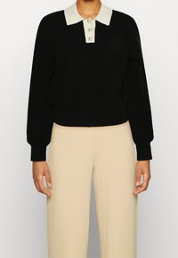 Pull noir en tricot avec un col crème à col et trois boutons décoratifs. Poignets ajustés et coupe décontractée.