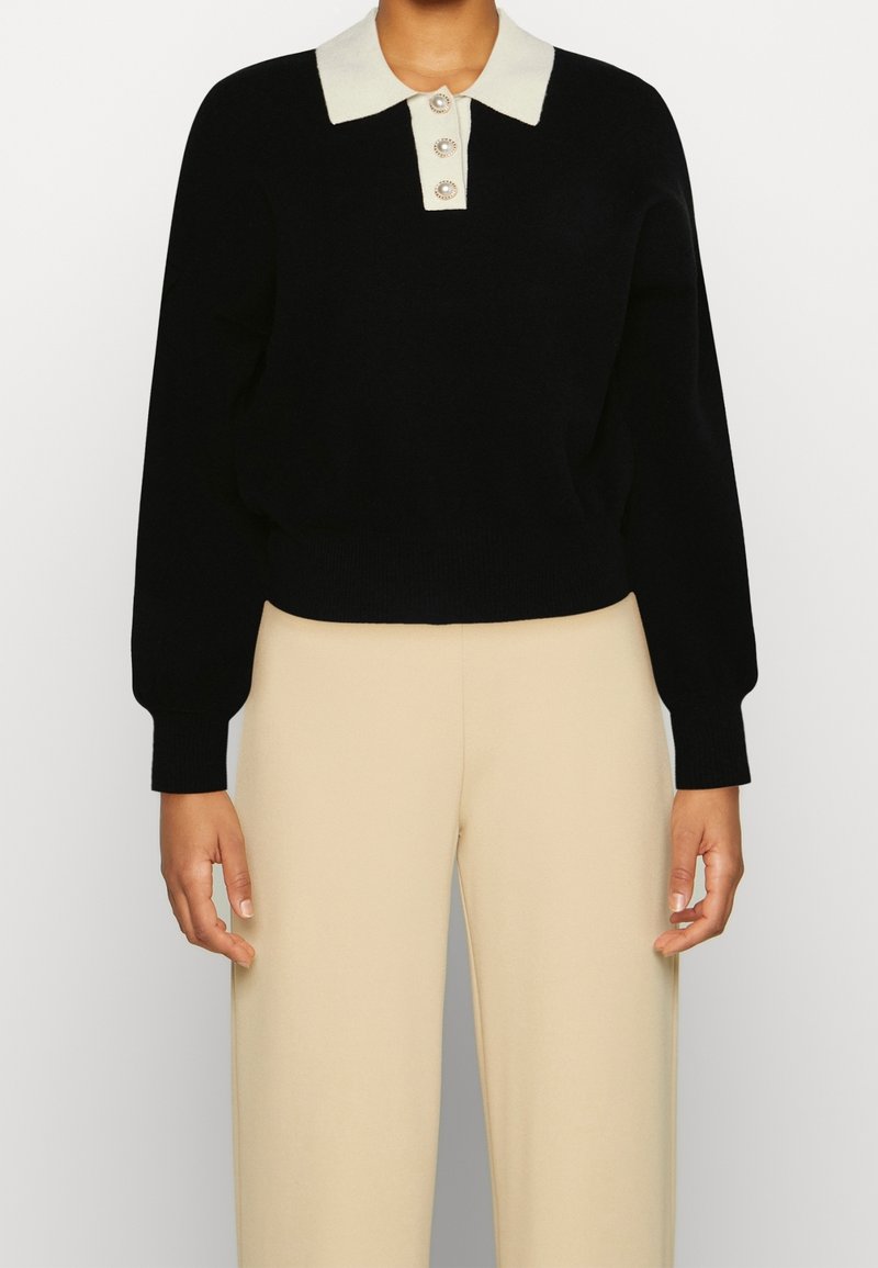 Pull noir en tricot avec un col crème à col et trois boutons décoratifs. Poignets ajustés et coupe décontractée.