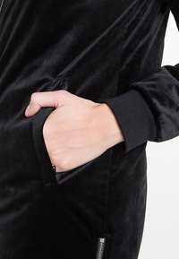 Juodas aksominis švarkas su užtrauktuko kišene ir banguotais rankogaliais. Tekstūra atrodo minkšta, turinti elegantišką dizainą ir minimalistiškus akcentus.