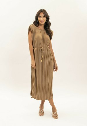 Femme aux longs cheveux noirs portant une robe midi plissée marron sans manches avec une ceinture nouée à la taille et des talons beiges à brides.