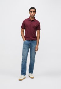 Bordowa koszulka polo z kołnierzykiem, noszona z jasnoniebieskimi dżinsami. Model stoi w swobodnej pozie, prezentując casualowe białe trampki z gumową podeszwą.