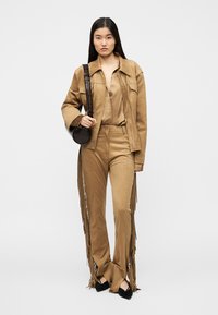 Μπεζ μπουφάν με φράντζες και παντελόνι που ταιριάζει, φτιαγμένα από μαλακό υλικό, συνδυασμένα με καφέ στρογγυλή τσάντα και μαύρα ψηλοτάκουνα· ομαλή υφή σε όλο το σύνολο.